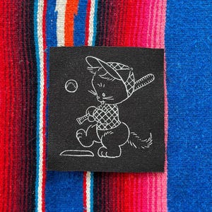May include: A square black fabric patch featuring a white line art illustration of a cartoon cat wearing a baseball cap and a plaid shirt, holding a baseball bat while standing at home plate with a baseball flying through the air nearby.