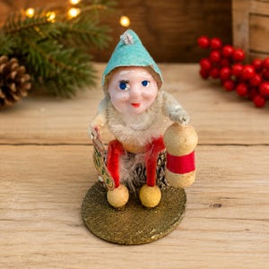 May include: A vintage-style gnome figurine with a light blue pointed hat, white fuzzy beard, and red legs. The gnome sits on a textured gold base, holding a small decorative stocking in one hand and a cylindrical beige object with a red band in the other.
