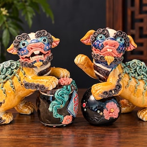 May include: A pair of ceramic Foo Dog statues featuring yellow bodies, dark blue and pink faces, and curly dark manes. One statue rests a paw on a black cylindrical drum, while the other rests a paw on a black sphere decorated with a pink flower.