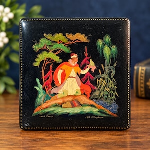 May include: A square black lacquer box featuring a hand-painted scene of a figure in a yellow tunic and red cape kneeling on a hill, holding a frog. The artwork includes green trees, red foliage, and the text 1975 B. Perekhin in the bottom corner.