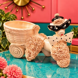 May include: A ceramic poodle figurine planter featuring a cream-colored basket on its back. The poodle has textured beige fur, wears a black beret, black cat-eye glasses, and a white bow tie with black polka dots. It has a red nose.