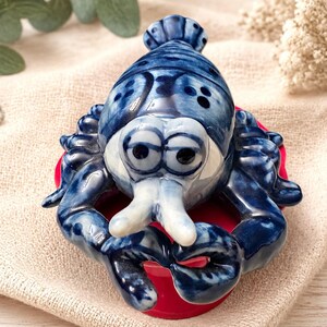 May include: A small, glossy ceramic figurine of a lobster featuring a deep blue and white marbled glaze. The lobster has large, rounded eyes, two prominent front claws, and a segmented tail, resting atop a circular, solid red base.