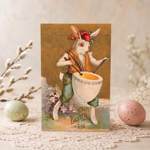 May include: A vintage-style postcard featuring an illustration of a white rabbit wearing a red cap and orange vest, playing a large white drum. The text "HAPPY EASTER TIDE" is printed at the bottom. The rabbit stands among small purple flowers.