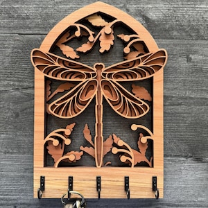 Può includere: Portachiavi in legno con un design a libellula e cinque ganci. La libellula è finemente intagliata e circondata da foglie. Il portachiavi è tinto di un colore marrone chiaro.
