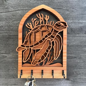 May include: Wooden key holder featuring a layered design of a sea turtle and coral. The key holder is made of light brown wood with a dark brown background and includes five metal hooks for keys.
