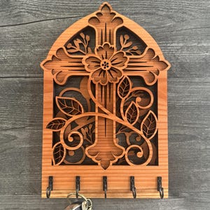 May include: Wooden key holder with a cross and floral design. The key holder is made of light brown wood and features a cutout cross with a flower and leaves. It has five metal hooks for keys.