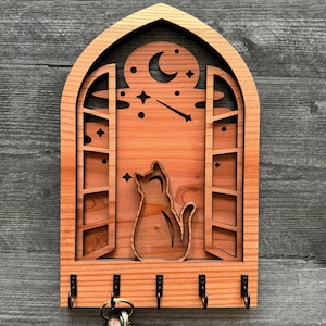 May include: Wooden key holder shaped like a window with a cat looking out at a crescent moon and stars. There are five hooks on the bottom for hanging keys.