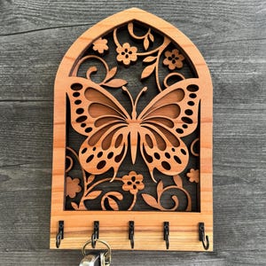 Puede incluir: Llavero de madera con diseño de mariposa. El llavero está hecho de madera de color marrón claro y presenta un patrón de mariposa y flores cortado con láser. Tiene cinco ganchos negros para llaves y una parte superior arqueada decorativa.