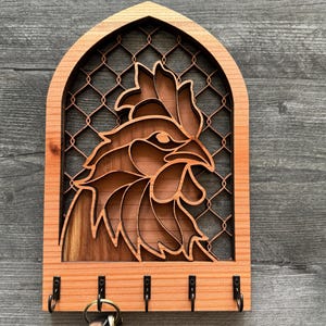 Könnte beinhalten: Schlüsselhalter aus Holz mit einem Hahn-Design. Der Hahn ist in das Holz geschnitzt und hat ein detailliertes Design. Der Schlüsselhalter hat fünf Haken zum Aufhängen von Schlüsseln.