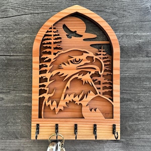 Könnte beinhalten: Schlüsselhalter aus Holz mit einer geschnitzten Darstellung eines Adlerkopfes in einer Waldlandschaft. Der Schlüsselhalter hat fünf Haken zum Aufhängen von Schlüsseln.