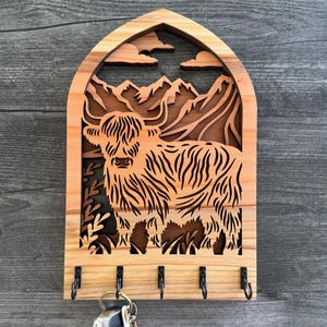 May include: Wooden key holder featuring a Highland cow design. The key holder is made of light brown wood and has a cutout design of a cow, mountains, and clouds. Includes five black metal hooks for keys.