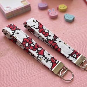 May include: Two fabric wristlet keychains featuring a repeating pattern of the Hello Kitty character on a white background. Each keychain has a silver-toned metal clasp and a circular split ring attached to one end for holding keys securely.