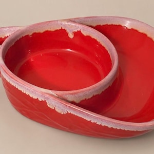 May include: A bright red ceramic serving dish with a glossy finish and a unique asymmetrical shape. The piece features a circular central bowl integrated into a larger, curved section, with a handle on one side and light pink trim.