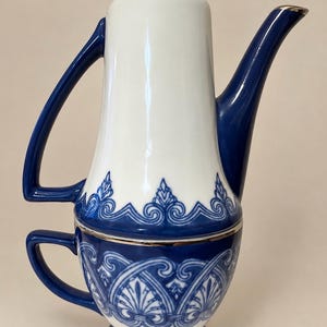 May include: A ceramic tea set for one featuring a white teapot stacked atop a matching blue and white teacup. The set is decorated with intricate blue scrollwork patterns and finished with metallic silver trim on the lid, spout, and rims.