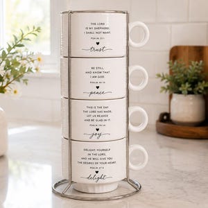 May include: A set of four white ceramic mugs stacked vertically in a silver metal wire stand. Each mug features black printed text with Bible verses and the words trust, peace, joy, and delight, accompanied by a small black heart icon below each verse.