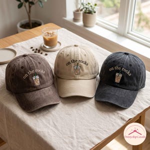 Puede incluir: Tres gorras de béisbol en marrón oscuro, beige y gris carbón. Cada gorra presenta un gráfico bordado de una bebida helada en un vaso de plástico transparente con una pajita verde y el texto "on the rocks" cosido en hilo negro.