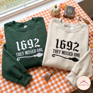 Peut inclure: Un sweat-shirt vert foncé avec une broderie blanche et un sweat-shirt crème avec une broderie noire. Les deux présentent le texte 1692 THEY MISSED ONE et un motif de balai. Les vêtements ont des manches longues, des poignets côtelés et un col rond.
