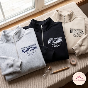 May include: Three quarter-zip sweatshirts in light gray, black, and beige. Each features the text CUSTOM UNIVERSITY NURSING with a stethoscope heart graphic. The gray sweatshirt has dark blue text, while the black and beige have white text.