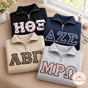 May include: Four quarter-zip sweatshirts in black, navy blue, beige, and light grey. Each features large Greek-style letters with distinct patterns: cherry print, white polka dots on navy, leopard print, and pink polka dots on a light background.