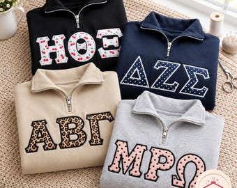 May include: Four quarter-zip sweatshirts in black, navy blue, beige, and light gray. Each features large Greek-style letters with distinct patterns: cherry print, white polka dots on navy, leopard print, and pink polka dots on a light background.