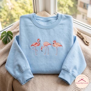 Könnte beinhalten: Ein hellblaues Sweatshirt mit Rundhalsausschnitt und einer Stickerei von drei rosafarbenen Flamingos in verschiedenen Posen auf der Brustmitte. Das Kleidungsstück hat lange Ärmel mit gerippten Bündchen sowie einen gerippten Halsausschnitt.