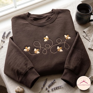 May include: A dark brown crewneck sweatshirt featuring an embroidered design of four yellow and white bees. The bees are connected by a series of stitched, dashed lines that create a looping, swirling pattern across the center of the chest.