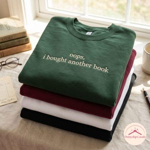 Puede incluir: Una pila de cuatro sudaderas de cuello redondo en verde oscuro, granate, blanco y negro. La sudadera verde oscuro superior presenta el texto blanco bordado "oops, i bought another book" centrado en el pecho. Cada sudadera tiene un cuello acanalado.