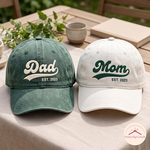 Puede incluir: Una gorra de béisbol verde oscuro con texto cursivo blanco que dice Dad EST. 2023 y una gorra blanca con texto cursivo verde oscuro que dice Mom EST. 2023. Ambas gorras tienen una corona estructurada, visera curva y letras bordadas al frente.