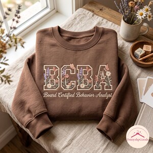 May include: A brown crewneck sweatshirt featuring the embroidered letters BCBA in a collegiate font, decorated with floral and butterfly motifs. Below the letters, the text Board Certified Behavior Analyst is printed in a white script font.