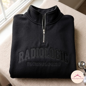 以下が含まれることがあります： 胸元にRADIOLOGIC TECHNOLOGISTの文字が同色の黒い糸で刺繍された、黒いクォータージップのプルオーバー。襟はハイネックで、中央にシルバーカラーの金属製ジッパーが付いています。