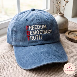 Puede incluir: Una gorra de béisbol de mezclilla azul con texto bordado en rojo y blanco. Las letras F, D y T están cosidas en rojo, mientras que las palabras FREEDOM, DEMOCRACY y TRUTH están cosidas en blanco, dispuestas verticalmente en el frente.