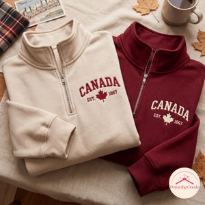 Könnte beinhalten: Zwei Sweatshirts mit kurzem Reißverschluss, eines in Creme und eines in Kastanienbraun, jeweils mit der Stickerei CANADA EST. 1867 und einem Ahornblatt-Symbol. Das cremefarbene Modell hat kastanienbraune Stickereien, das kastanienbraune Modell cremefarbene.