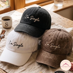 Puede incluir: Tres gorras de béisbol en negro, blanco y marrón, cada una con el texto bordado I'm Single en una fuente cursiva. Las gorras tienen una corona estructurada, una visera curva y un botón en la parte superior. Las gorras negra y marrón tienen un acabado desgastado.
