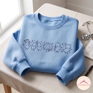 Puede incluir: Una sudadera de cuello redondo en color azul claro con un estampado gráfico en azul oscuro de cinco órganos humanos tipo dibujos animados, incluyendo corazón, pulmones, cerebro, riñón y estómago, cada uno con cara sonriente y extremidades.