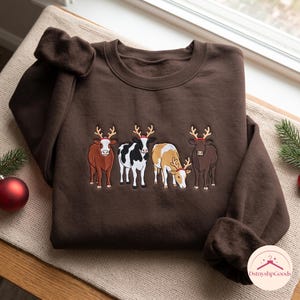 Könnte beinhalten: Ein dunkelbraunes Sweatshirt mit Rundhalsausschnitt und einer Stickerei von vier Kühen, die rote Rentiergeweihe tragen. Die Kühe sind in verschiedenen Farben dargestellt, darunter einfarbig braun, schwarz-weiß gefleckt, beige-weiß und dunkelbraun.