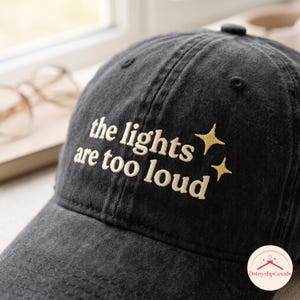 Könnte beinhalten: Eine schwarze Baseballkappe mit verwaschener Textur und cremefarbenem Sticktext, der the lights are too loud lautet, ergänzt durch zwei kleine vierzackige Sternsymbole, die mit passendem cremefarbenem Garn auf der rechten Seite gestickt sind.