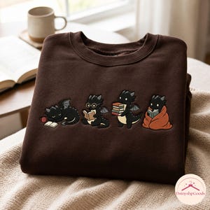 May include: A dark brown crewneck sweatshirt featuring an embroidered graphic of four small black dragons. The dragons are depicted in various activities: sleeping by a book, reading with glasses, holding a stack of books, and wrapped in a blanket.