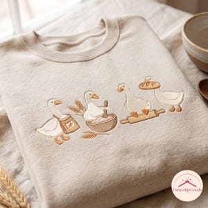 Könnte beinhalten: Ein beigefarbenes Sweatshirt mit Rundhalsausschnitt und einer Stickerei von vier weißen Gänsen beim Backen. Die Gänse sind dargestellt, wie sie einen Mehlsack tragen, Teig in einer Schüssel rühren, ein Nudelholz benutzen und ein Brot tragen.