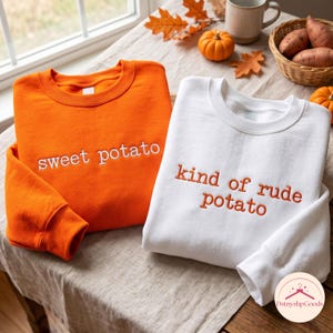 May include: An orange sweatshirt with white embroidered text reading sweet potato and a white sweatshirt with orange embroidered text reading kind of rude potato. Both garments feature a crew neckline and long sleeves with ribbed cuffs.