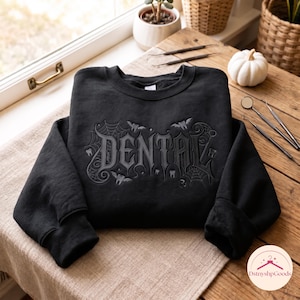 Op de afbeelding: Een zwart sweatshirt met ronde hals en donkergrijze borduursels van het woord DENTAL in een gotisch lettertype, omringd door spinnenwebben, kleine vleermuizen en tandiconen. Het ontwerp is gecentreerd op de borst van het kledingstuk met lange mouwen.