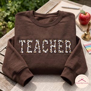 Könnte beinhalten: Ein dunkelbraunes Sweatshirt mit Rundhalsausschnitt und dem Wort TEACHER, das in Blockschrift auf die Brust gestickt ist. Jeder Buchstabe ist mit einem beigen, schwarzen und braunen Leopardenmuster gefüllt und dunkelbraun umrandet.