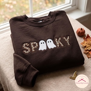 Könnte beinhalten: Ein dunkelbraunes Sweatshirt mit Rundhalsausschnitt und dem aufgestickten Wort SPOOKY auf der Brust. Die Buchstaben S, P, K und Y sind mit einem beigefarbenen und braunen Leopardenmuster gefüllt, während die beiden O durch weiße Geisterformen ersetzt sind.