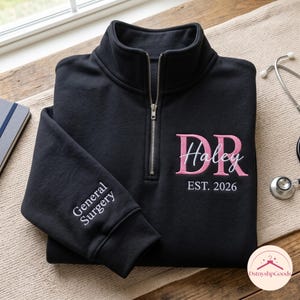 May include: A black quarter-zip sweatshirt featuring embroidered text. The front displays pink letters DR, white script text Haley, and white text EST. 2026. The sleeve is embroidered with white text reading General Surgery.
