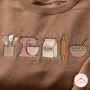 May include: A brown crewneck sweatshirt featuring embroidered baking-themed graphics. The design includes a utensil holder with a whisk, spatula, and spoon, a pink stand mixer, a bag labeled FLOUR, a wooden rolling pin, and a mixing bowl with a spoon.