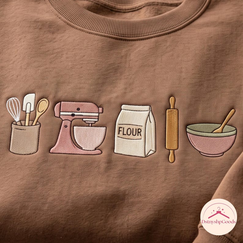 May include: A brown crewneck sweatshirt featuring embroidered baking-themed graphics. The design includes a utensil holder with a whisk, spatula, and spoon, a pink stand mixer, a bag labeled FLOUR, a wooden rolling pin, and a mixing bowl with a spoon.