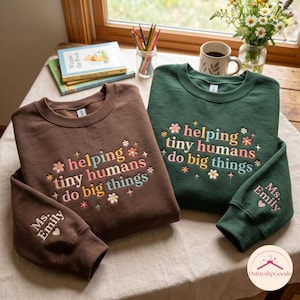 Peut inclure: Un sweat-shirt à col rond marron et un autre vert foncé, chacun arborant le texte brodé "helping tiny humans do big things" entouré de petites icônes de fleurs et d'étoiles. La manche de chaque sweat-shirt est brodée avec "Ms. Emily".