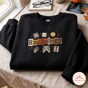 以下が含まれることがあります： マルチカラーのブロックフォントで「Radiology」という文字がプリントされた黒のクルーネックトレーナー。胸郭、頭蓋骨、手の骨格、長い骨、放射線マークのグラフィックイラストが描かれており、小さな星のアクセントが添えられています。