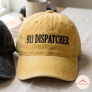 May include: A mustard yellow baseball cap with a distressed finish, featuring the text 911 DISPATCHER embroidered in black thread across the front panel. The cap has a structured crown, a curved brim, and visible stitching details throughout the fabric.