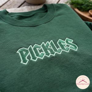 Op de afbeelding: Een bosgroene sweater met ronde hals en het woord PICKLES geborduurd op de borst in een vet, blokachtig lettertype. De letters zijn gestikt in een donkerdere tint groen met een licht mintgroene omlijning voor een gelaagd effect.