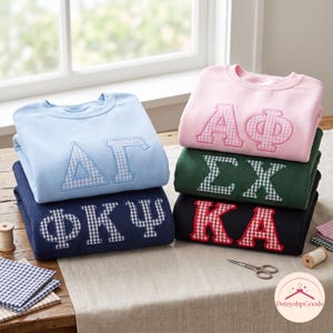 Peut inclure: Une pile de cinq sweat-shirts à col rond en bleu clair, bleu marine, rose clair, vert foncé et noir. Chaque sweat-shirt présente de grandes lettres grecques appliquées avec un motif à carreaux vichy aux couleurs assorties sur le devant.